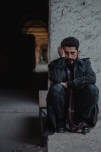 Despaired Ethnic Man In Dark Clothes Touching Head And Sitting On Stone Window Opening In Shabby Dusty Building